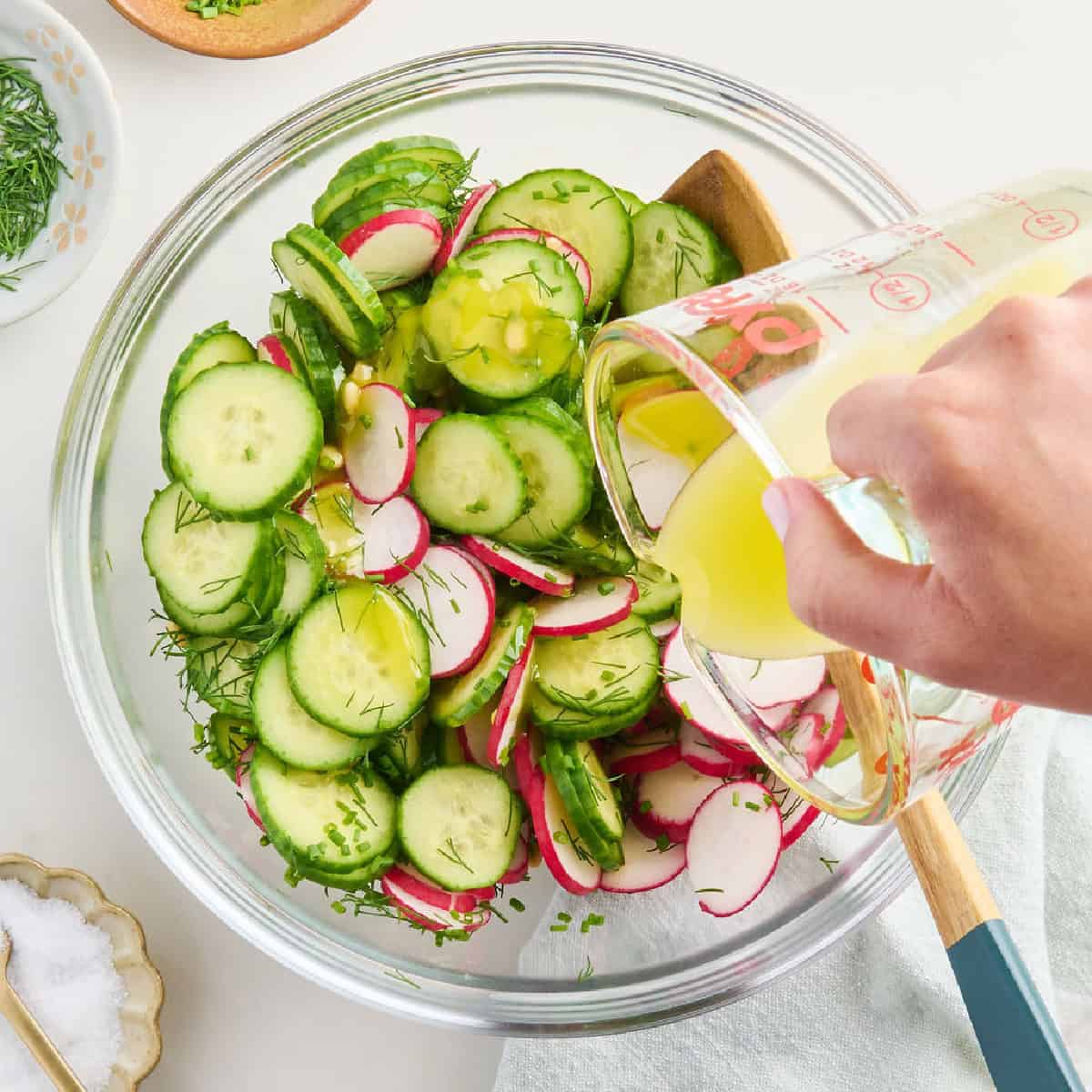 Woman pouring lemon dressing over a bowl of cucumber radish salad ingredients.