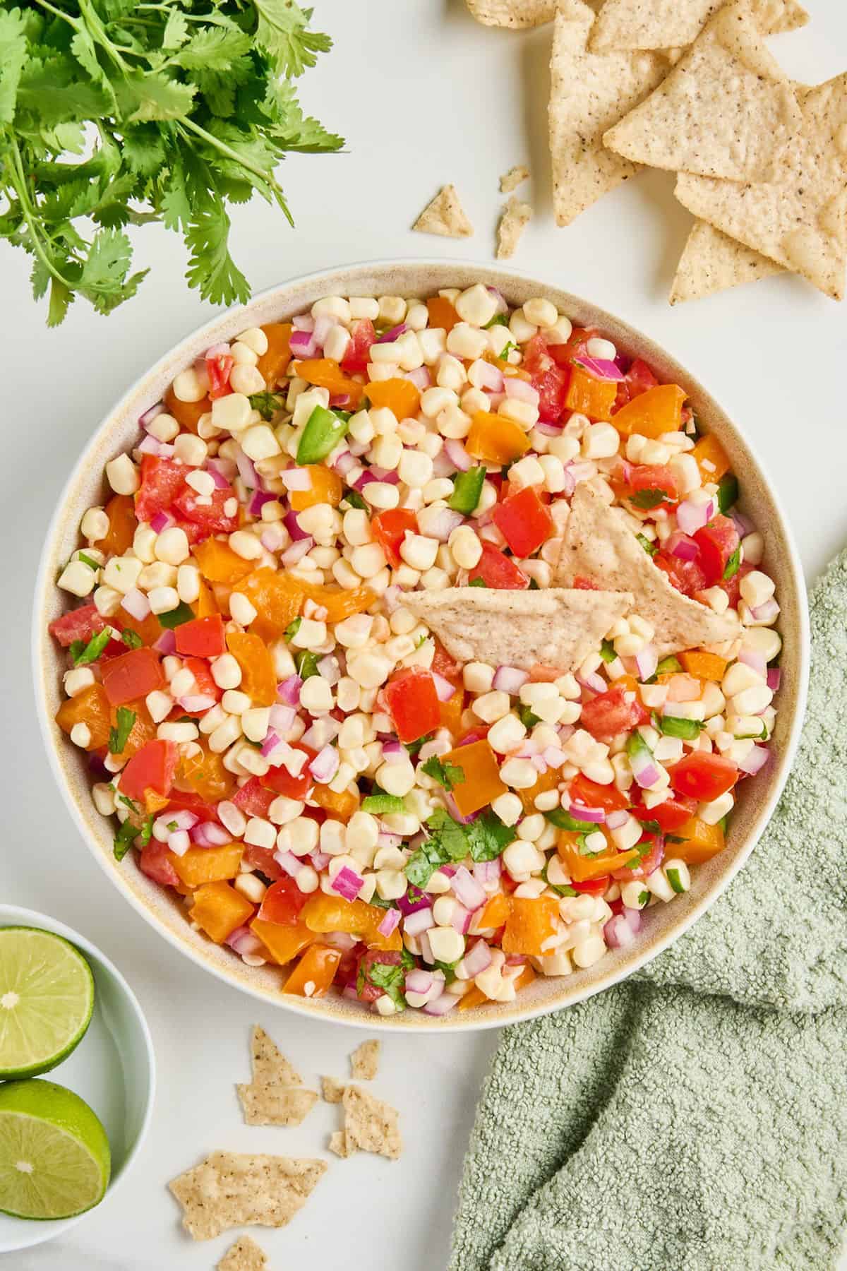 Corn salsa with two tortilla chips on top in a bowl next to ingredients and napkin.