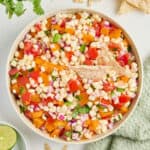 Corn salsa with two tortilla chips on top in a bowl next to ingredients and napkin.