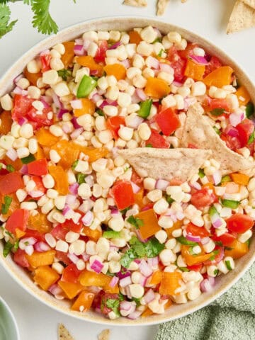 Corn salsa with two tortilla chips on top in a bowl next to ingredients and napkin.