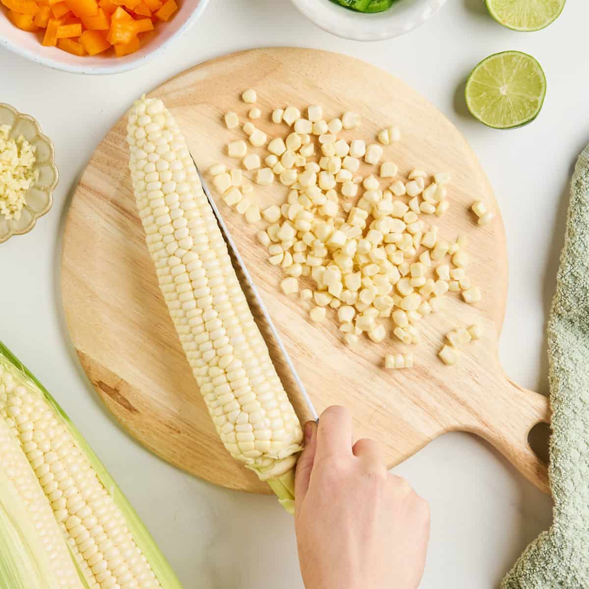 Woman slicing kernels off a corn cob with a serrated knife on a cutting board.
