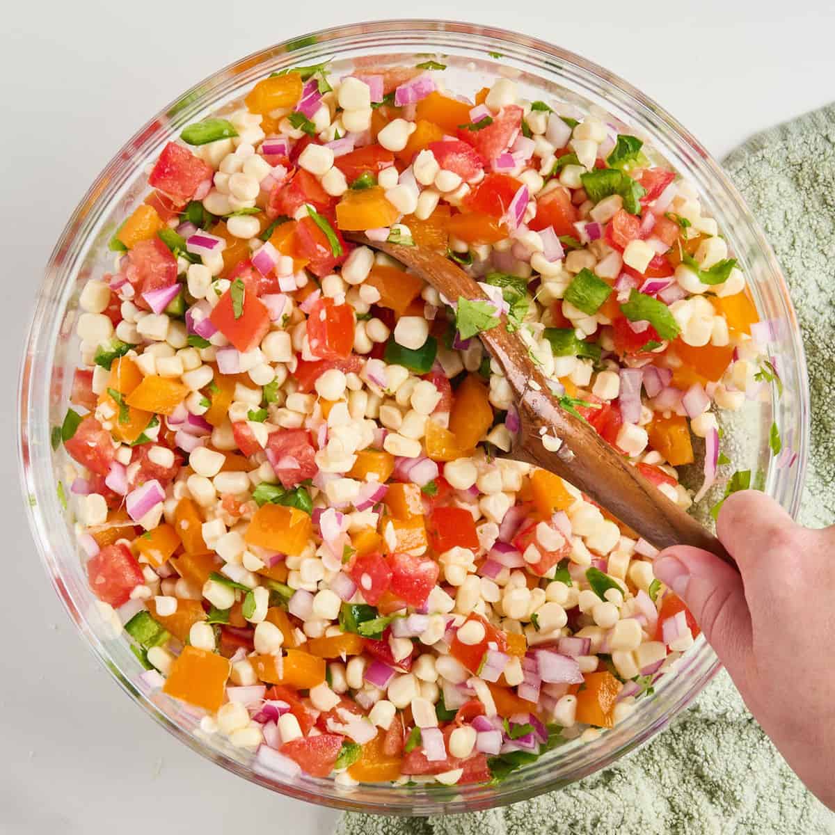 Woman stirring fresh corn and tomato salsa with a wooden spoon in a large bowl.