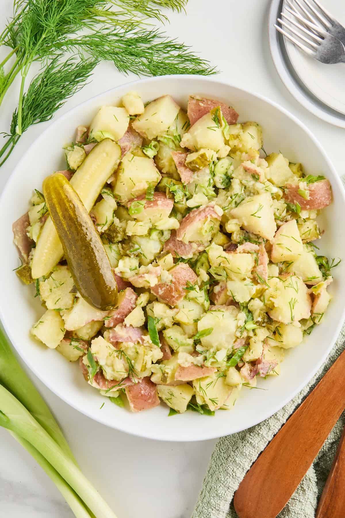 Dill pickle potato salad in a serving bowl next to ingredients, napkin, and plate with fork.