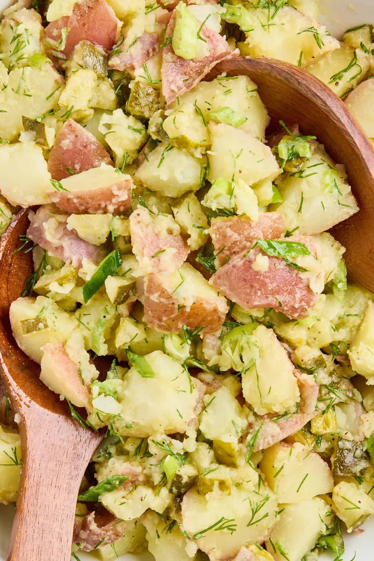 Potato salad with dill pickles and olive oil dressing being scooped with tongs.