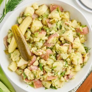 Dill pickle potato salad in a serving bowl next to ingredients, napkin, and plate with fork.