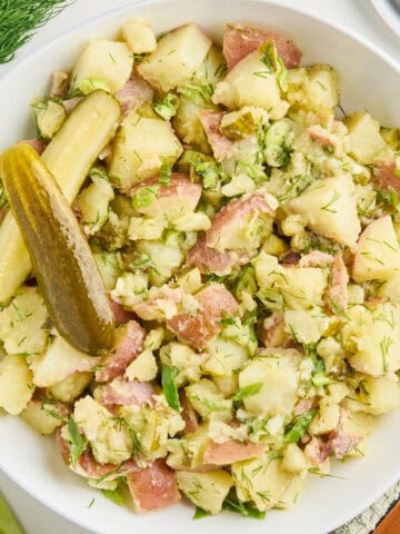 Dill pickle potato salad in a serving bowl next to ingredients, napkin, and plate with fork.