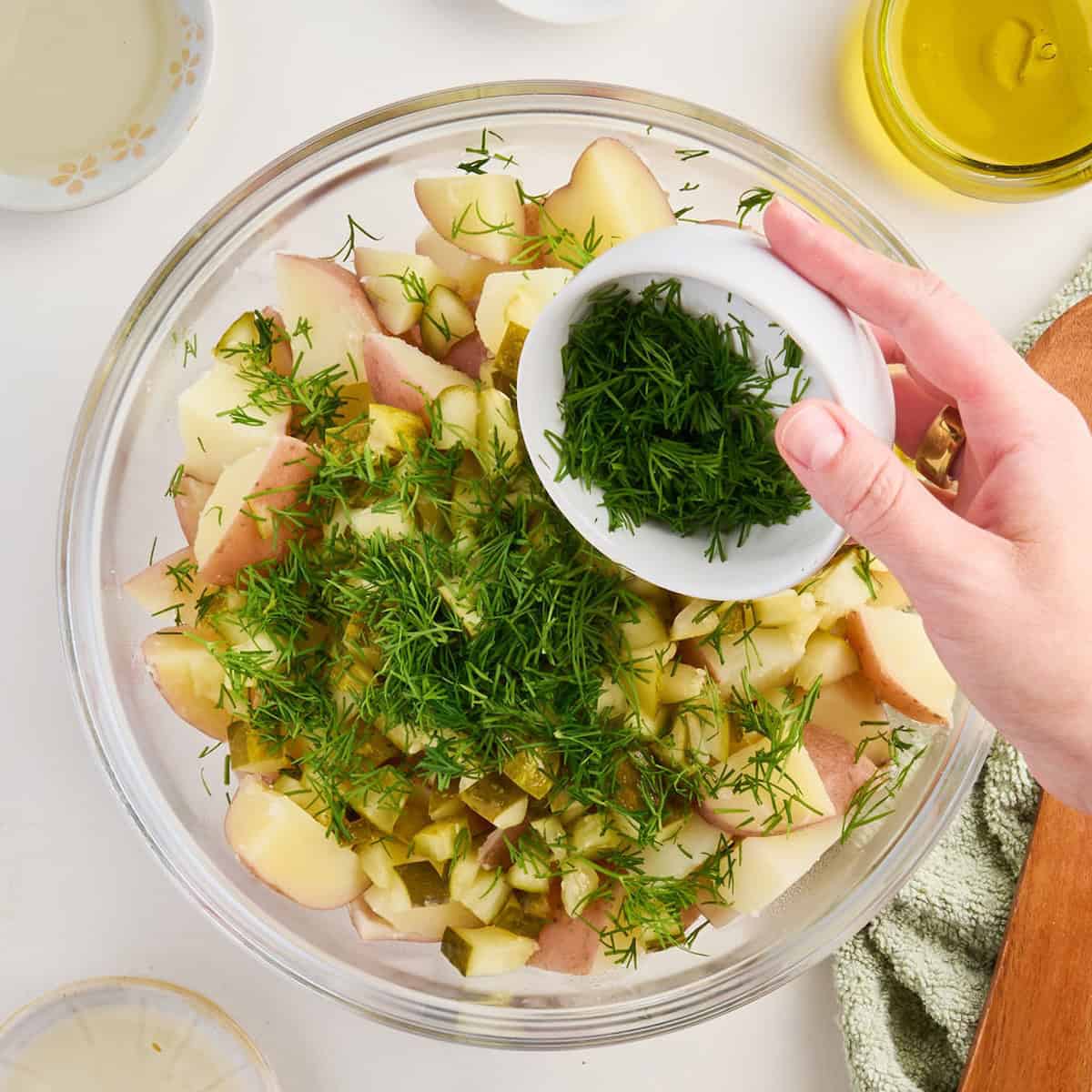 Woman adding chopped fresh dill to a bowl with potato salad ingredients.