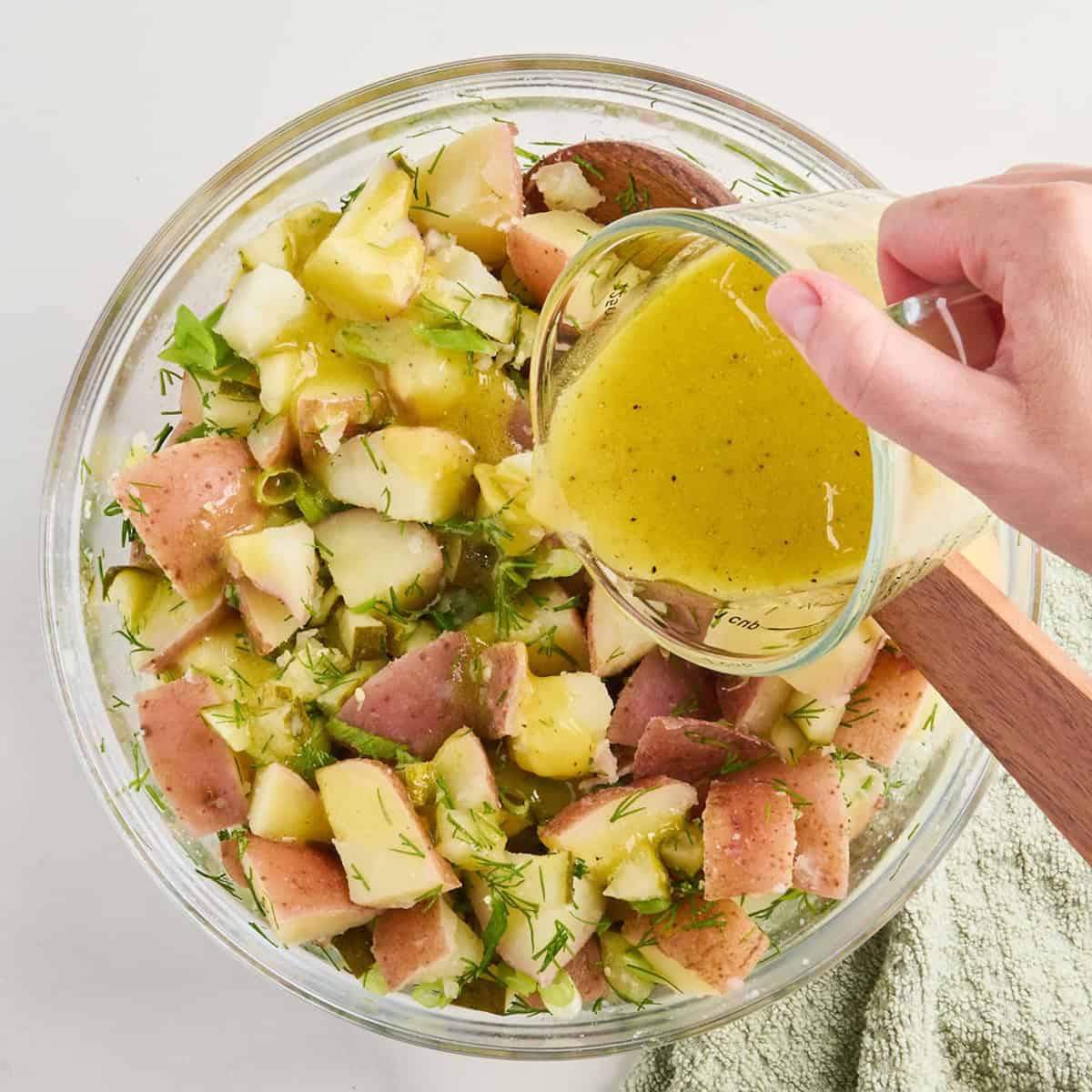 Woman pouring oil dressing over ingredients for potato salad in a bowl.