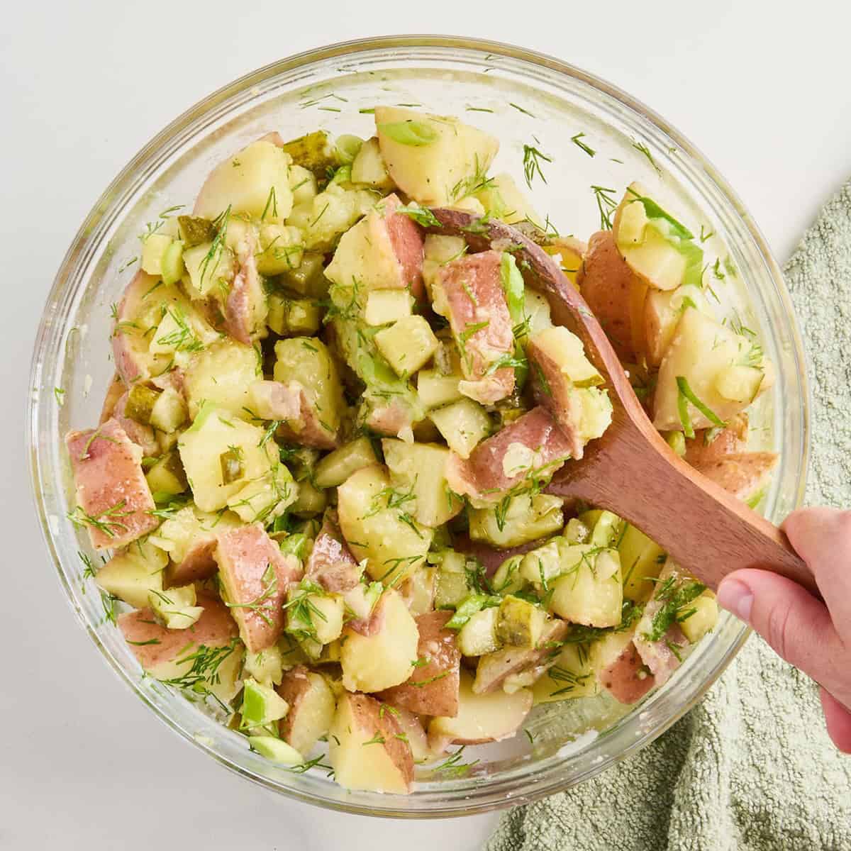 Woman mixing dill pickle potato salad with a spoon in a glass bowl.