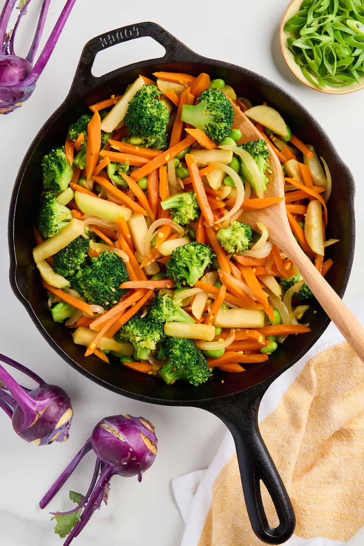 Kohlrabi stir fry in a cast iron skillet surrounded by purple kohlrabi, a napkin, and scallions.