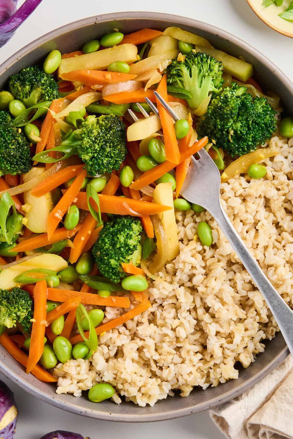 Stir fry with kohlrabi, broccoli, carrots, edamame, and brown rice with a fork on a plate.