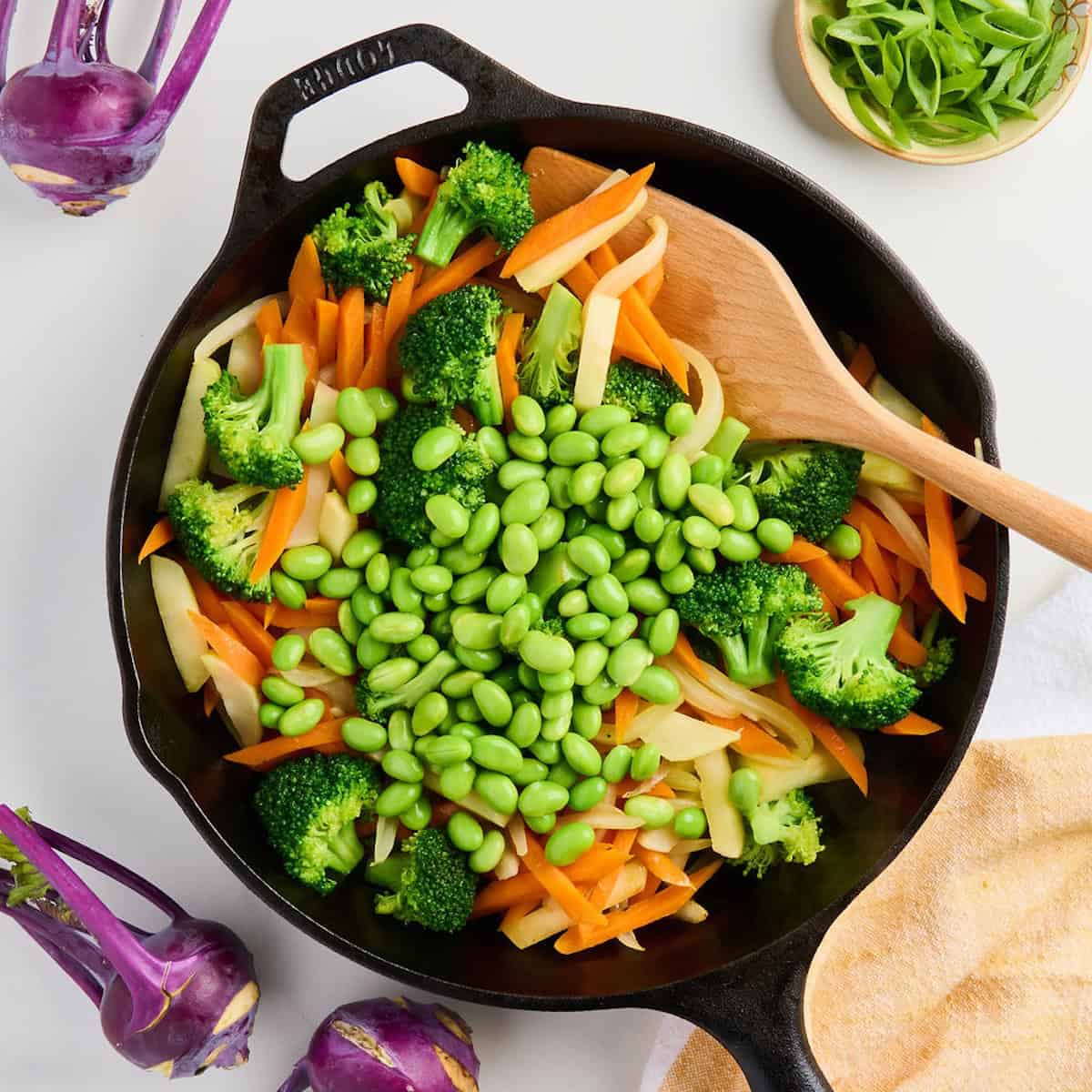 Shelled edamame added to a skillet with stir fry vegetables and a wooden spoon.
