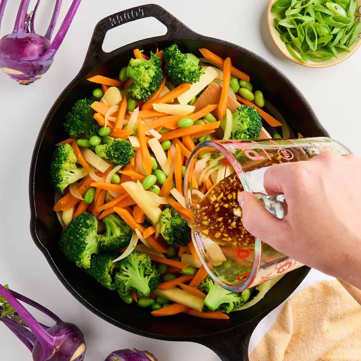 Woman pouring stir fry sauce over cooked vegetables and edamame in a skillet.