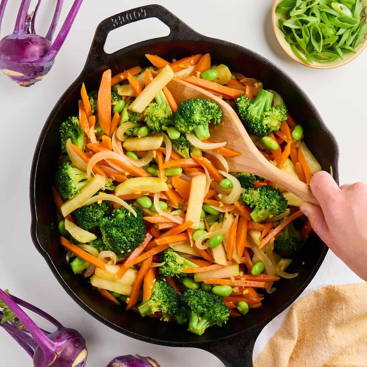 Woman stirring kohlrabi stir fry to coat it in sauce in a cast iron skillet.