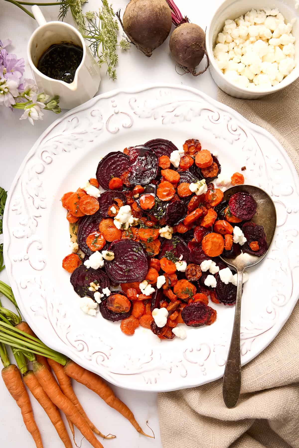 Roasted beets and carrots with goat cheese on a serving plate with spoon next to ingredients.