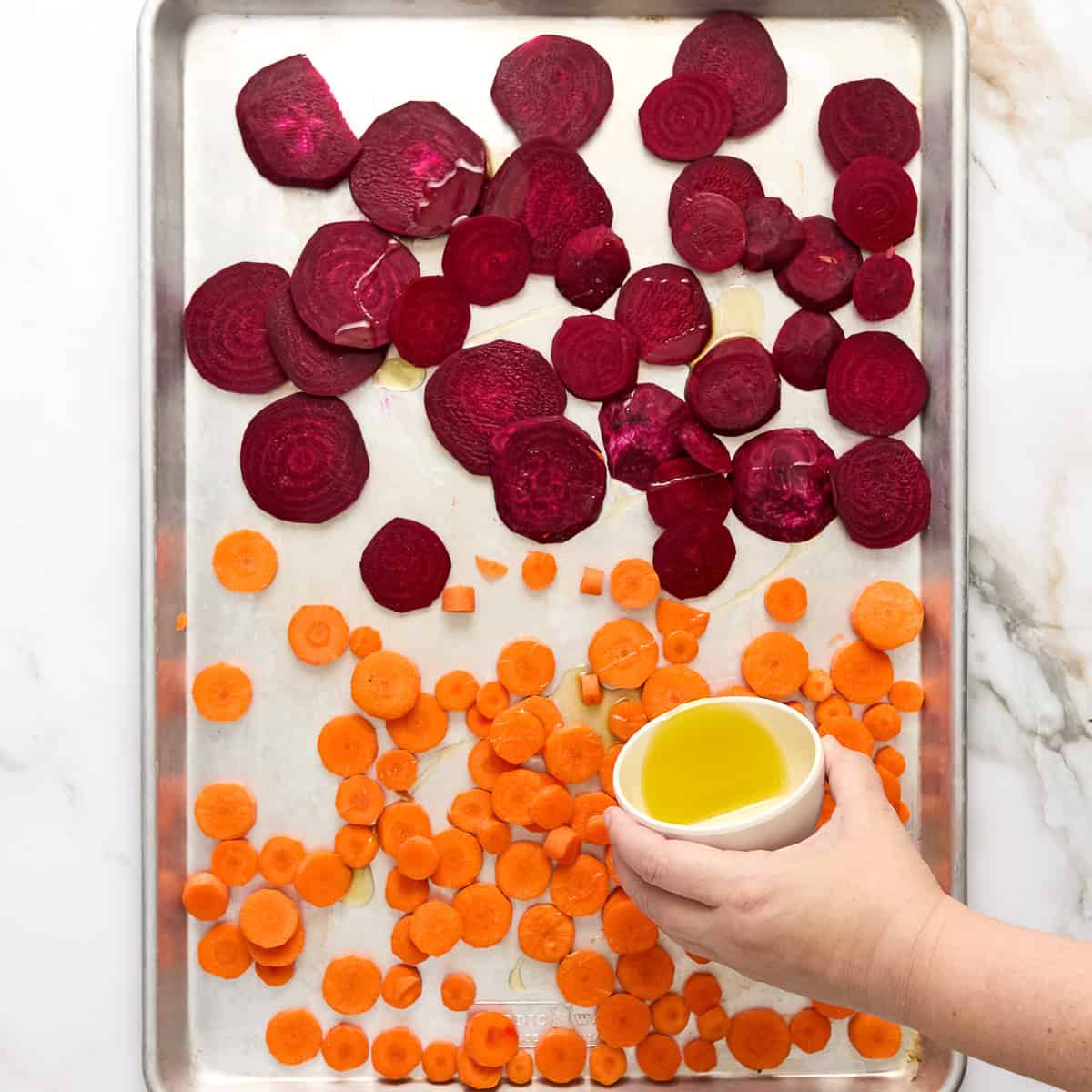 Woman pouring olive oil over sliced beets and carrots on a baking sheet.