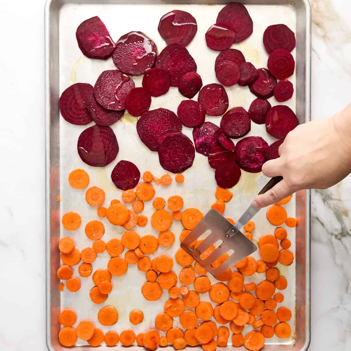 Woman tossing sliced beets and carrots in olive oil on a baking sheet.