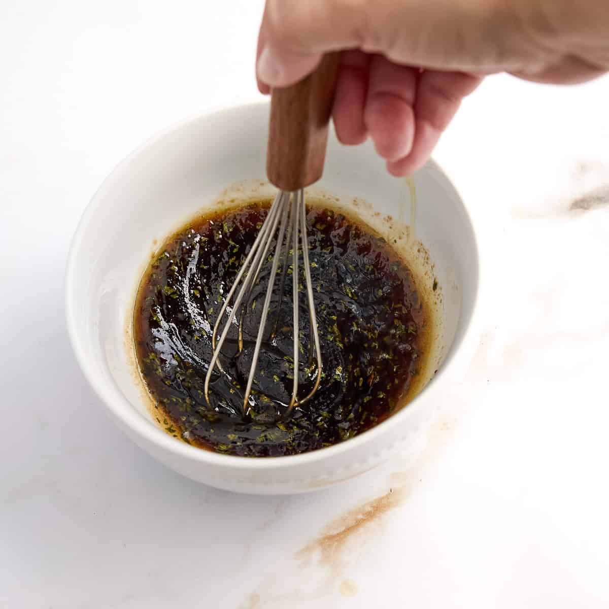 Woman whisking balsamic honey glaze with parsley in a small white bowl.