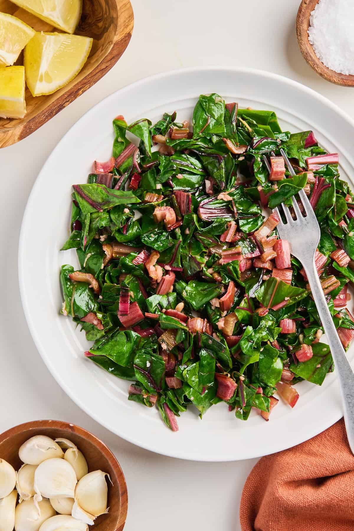 Cooked red chard with a fork next to lemons, garlic, salt, and napkin.