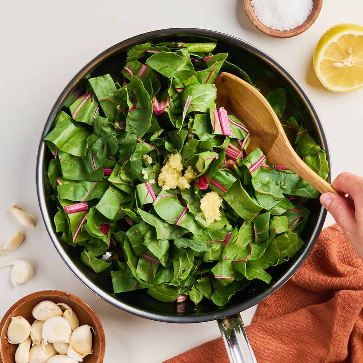 Woman stirring red chard leaves and minced garlic into a skillet.