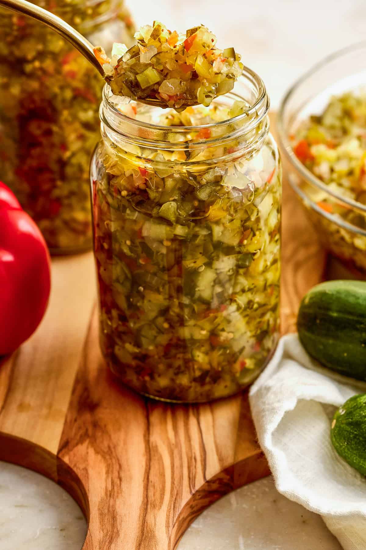 A spoonful of sweet relish being scooped out of a glass jar surrounded by ingredients.