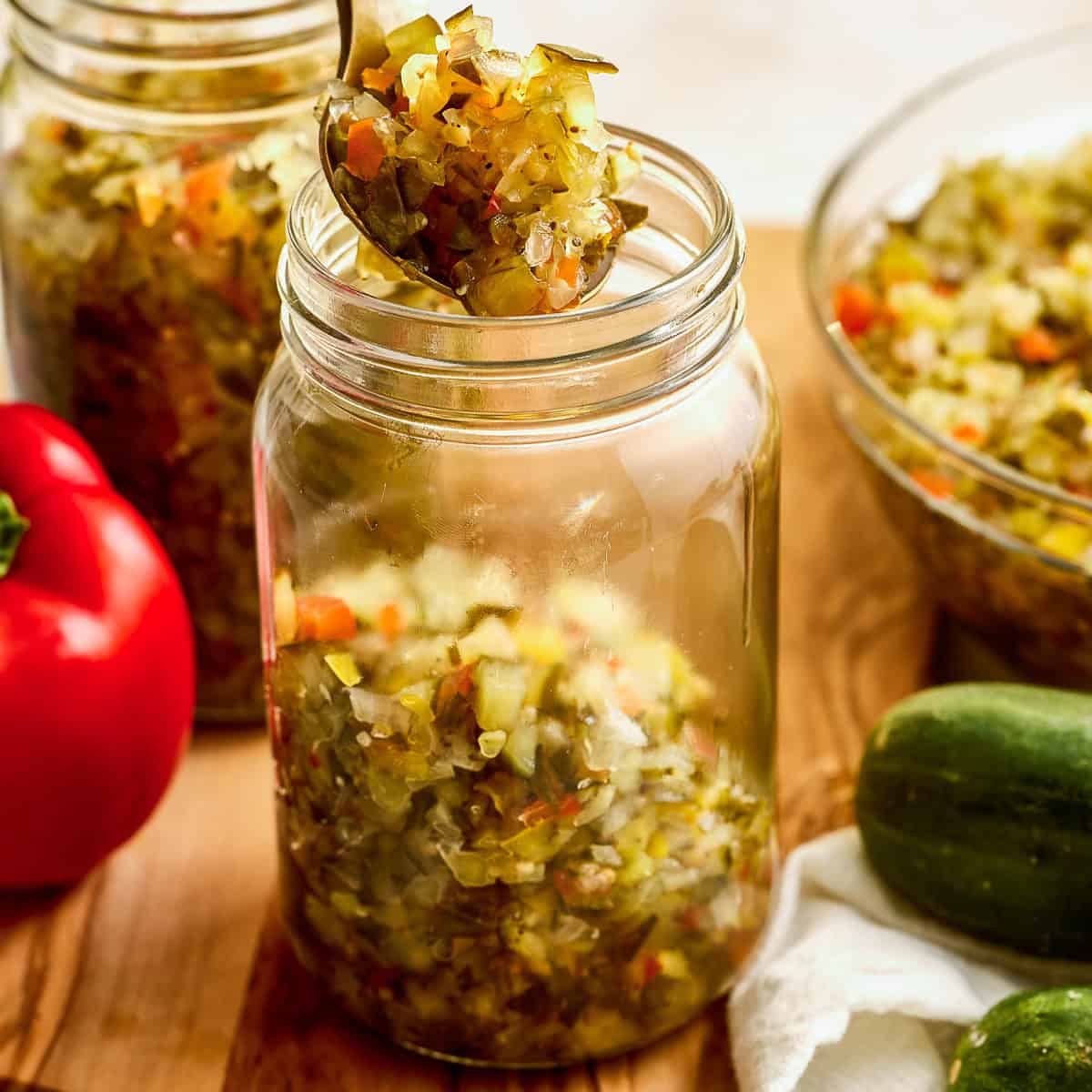 Sweet relish being spooned into a glass jar on a cutting board after cooking.