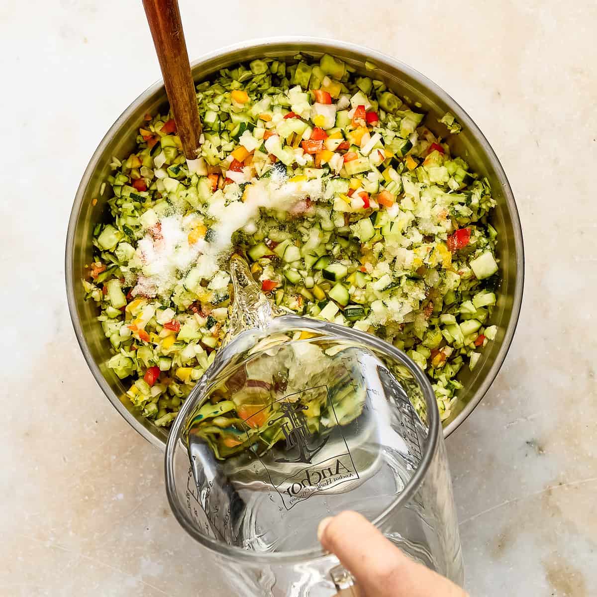 Woman pouring water into a bowl of cucumbers, peppers, onions, and kosher salt.