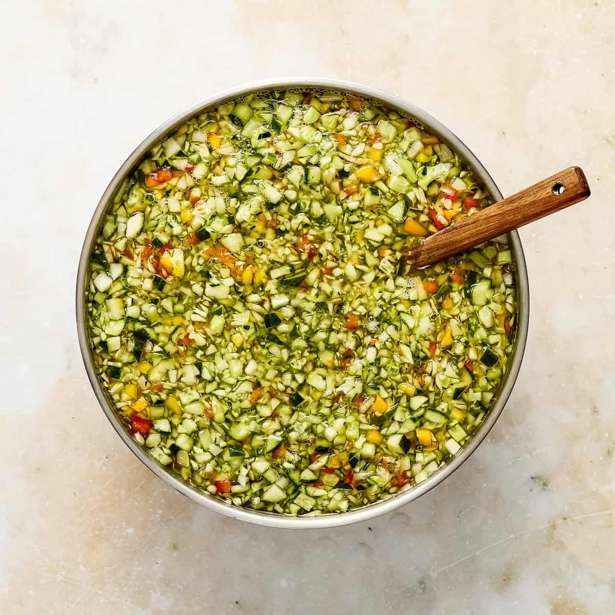 Cucumbers, peppers, and onions soaking in water and salt in a bowl with a spoon.