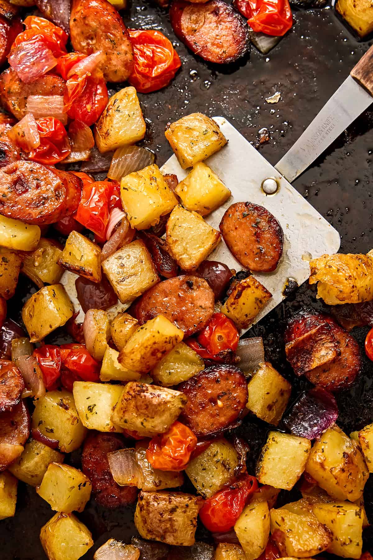 Potatoes, tomatoes, onion, and chicken sausage scooped with a spatula on a baking sheet.
