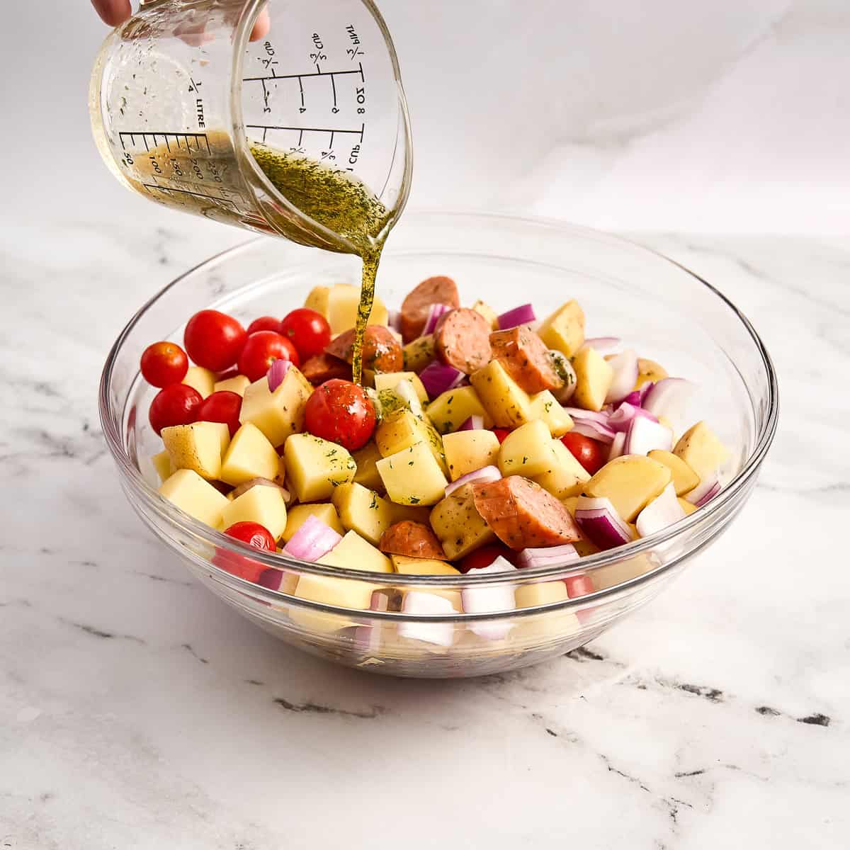 Woman pouring olive oil mixed with seasonings over chicken sausage and veggies in a bowl.