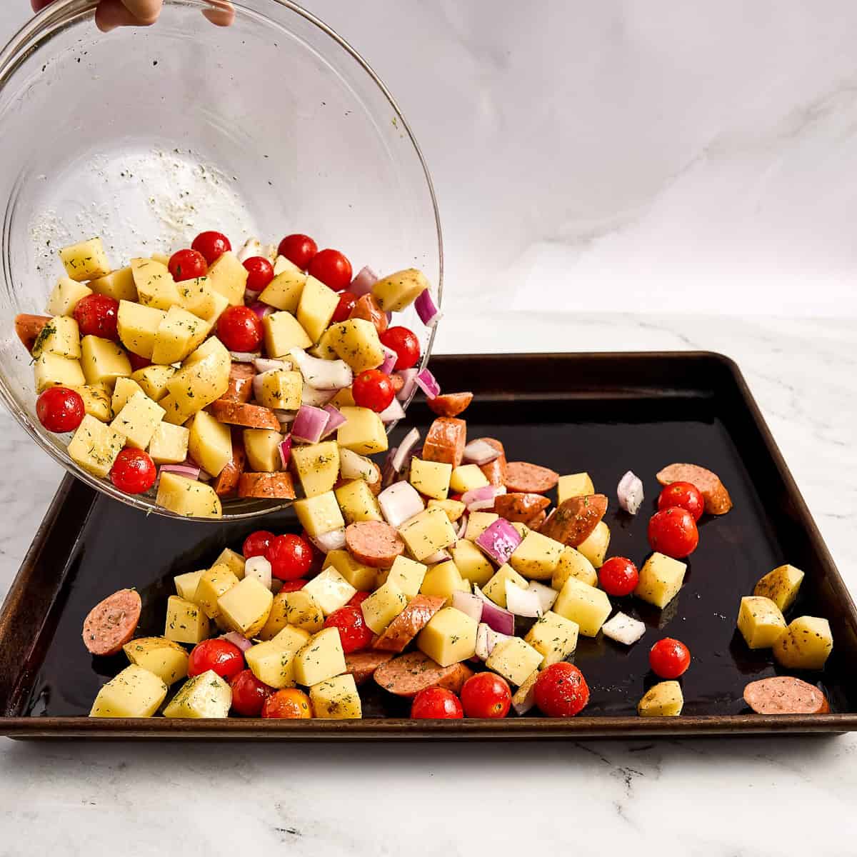 Woman pouring seasoned chicken sausage and veggies from a bowl onto a sheet pan.