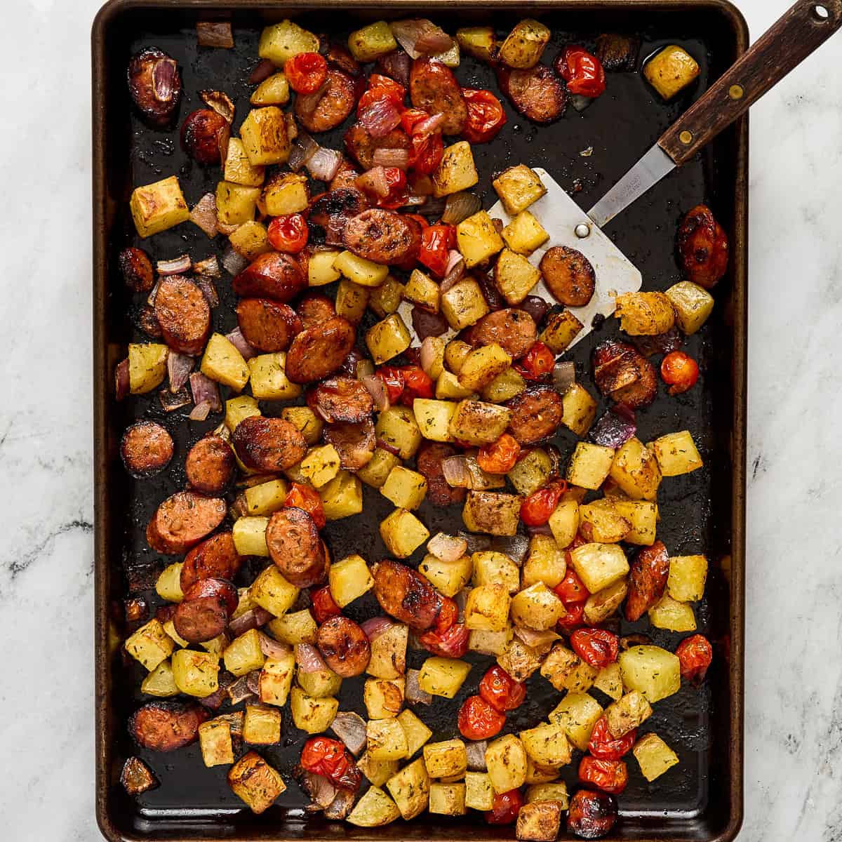 Roasted chicken sausage and veggies on a baking sheet with a metal spatula.