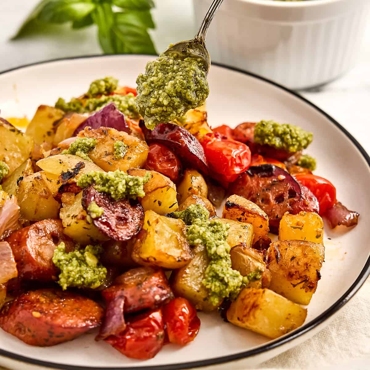 Woman spooning fresh pesto on top of a chicken sausage sheet pan dinner.