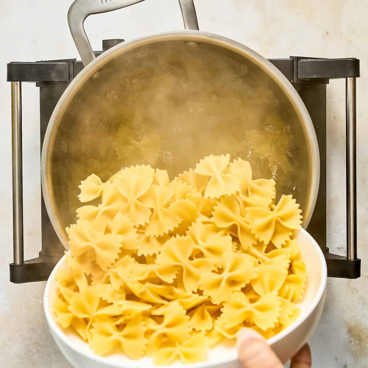 Bowtie pasta being poured into a pot of boiling water on a stove burner.