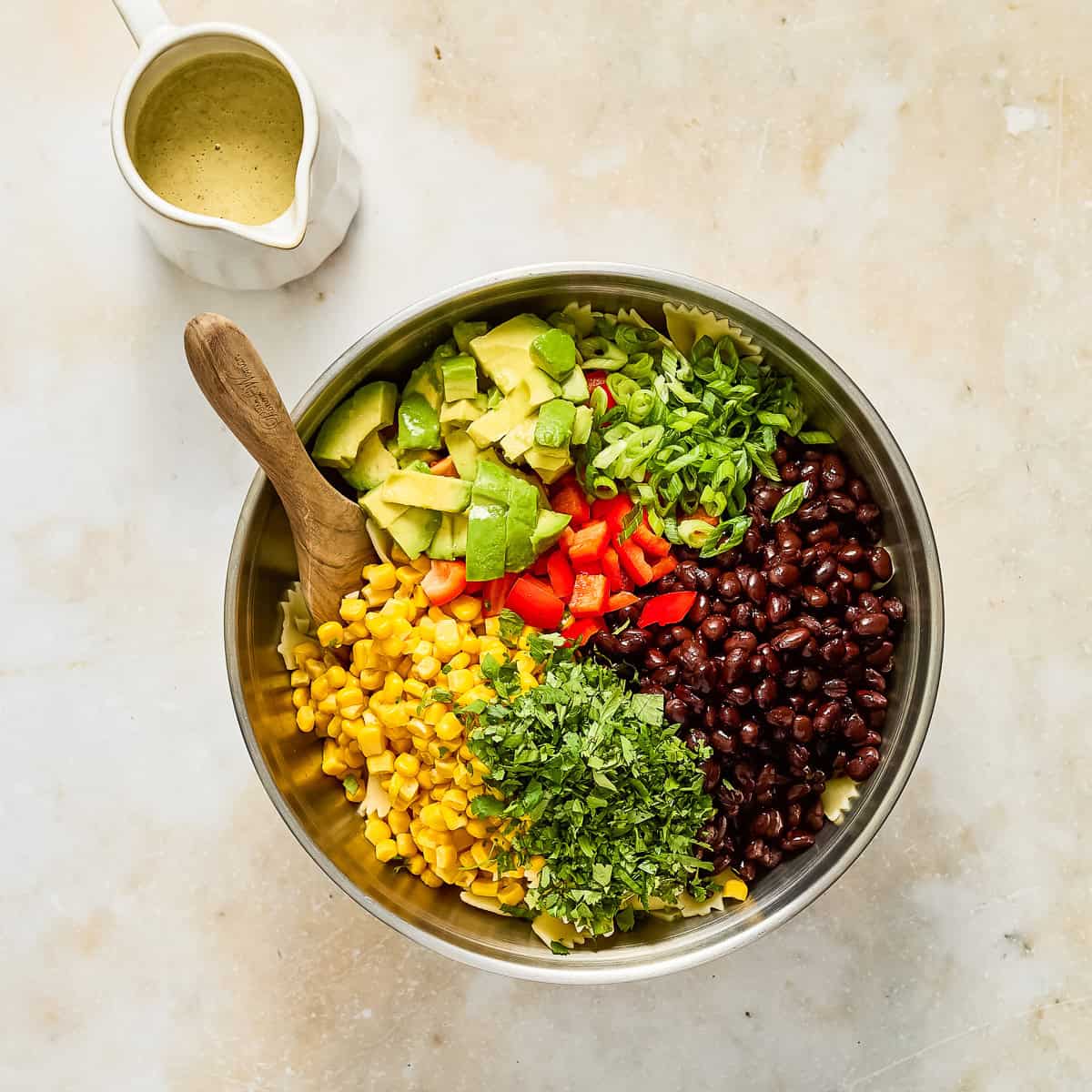 Pasta, corn, beans, avocado, pepper, cilantro, and scallions in a bowl next to pitcher of dressing.