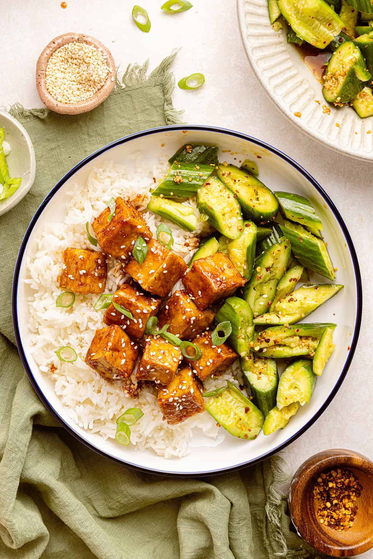 Sticky tofu with rice and cucumber salad surrounded by ingredients and toppings.