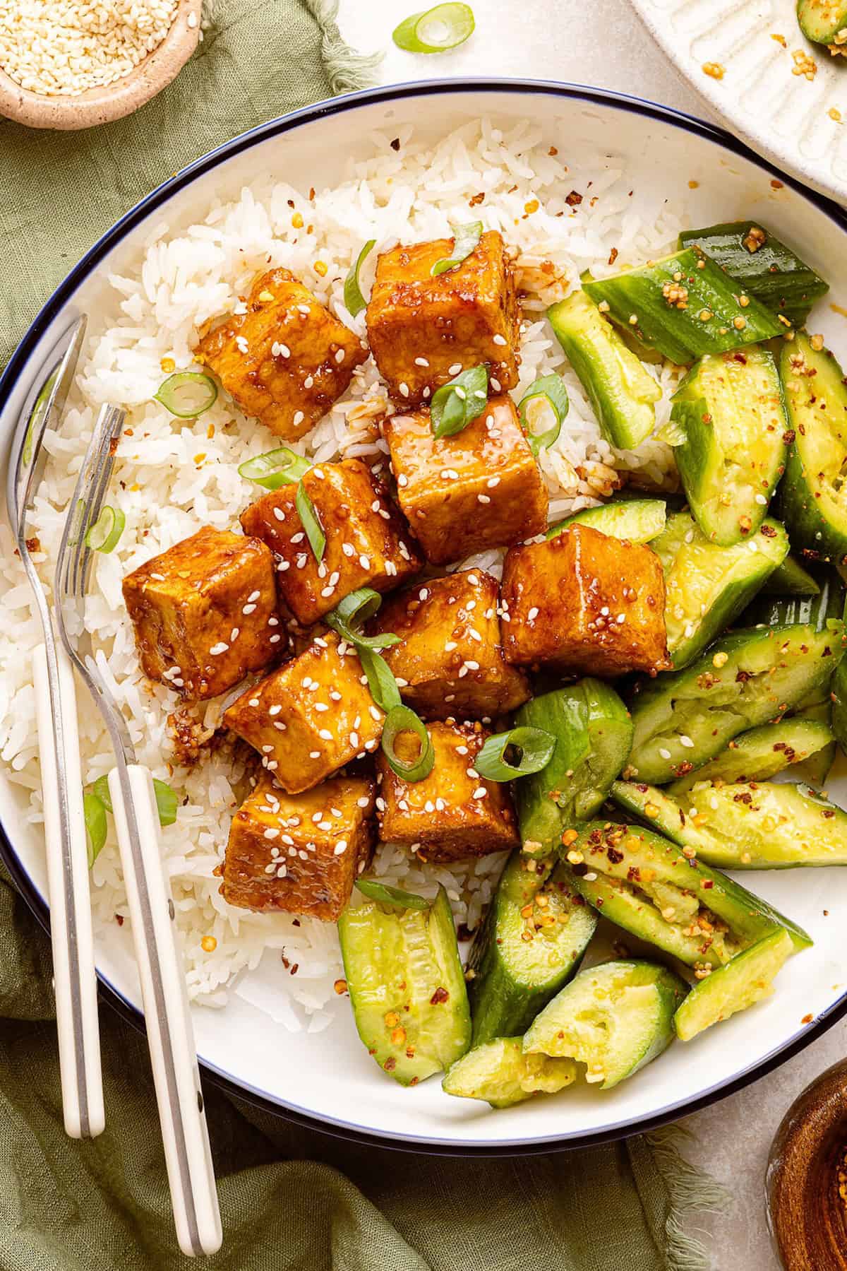 Bowl of sticky glazed tofu, white rice, and smashed cucumber salad with a fork and spoon.