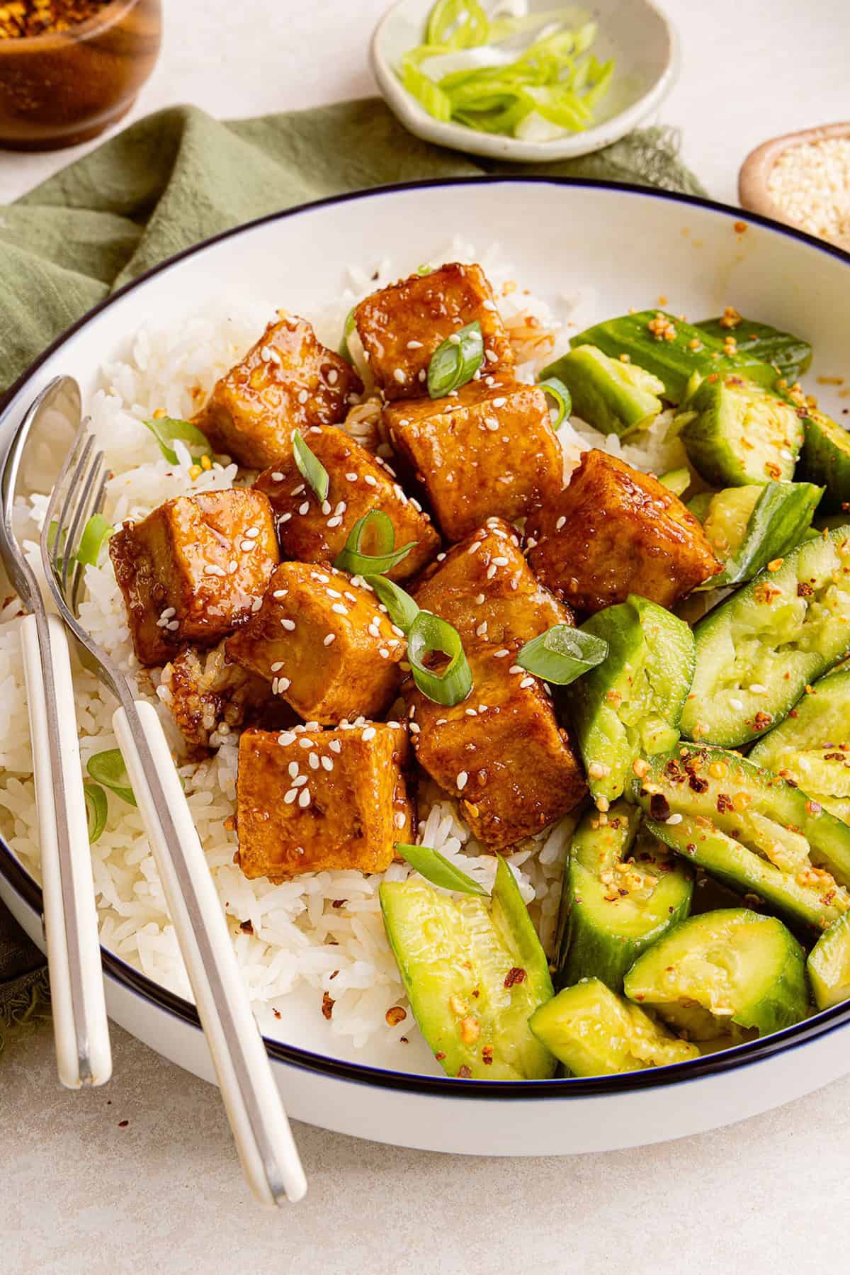 Tofu in a sweet sticky sauce served with rice and smashed cucumber salad in a bowl.