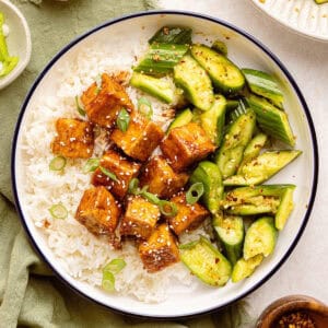 Sticky tofu with rice and cucumber salad surrounded by ingredients and toppings.