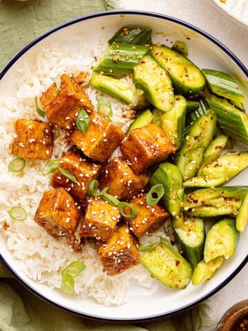 Sticky tofu with rice and cucumber salad surrounded by ingredients and toppings.