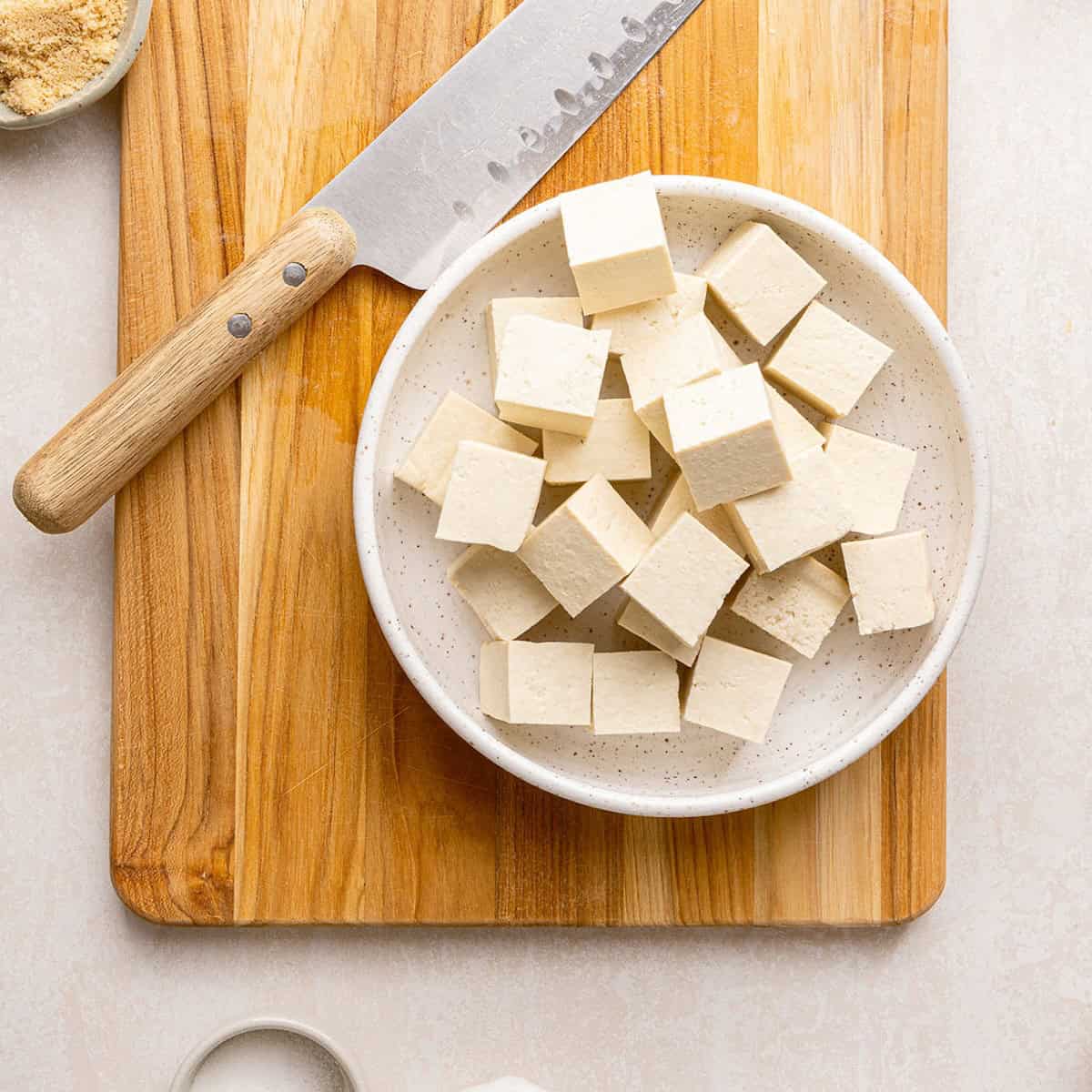Tofu sliced into cubes in a bowl on a cutting board with a knife.