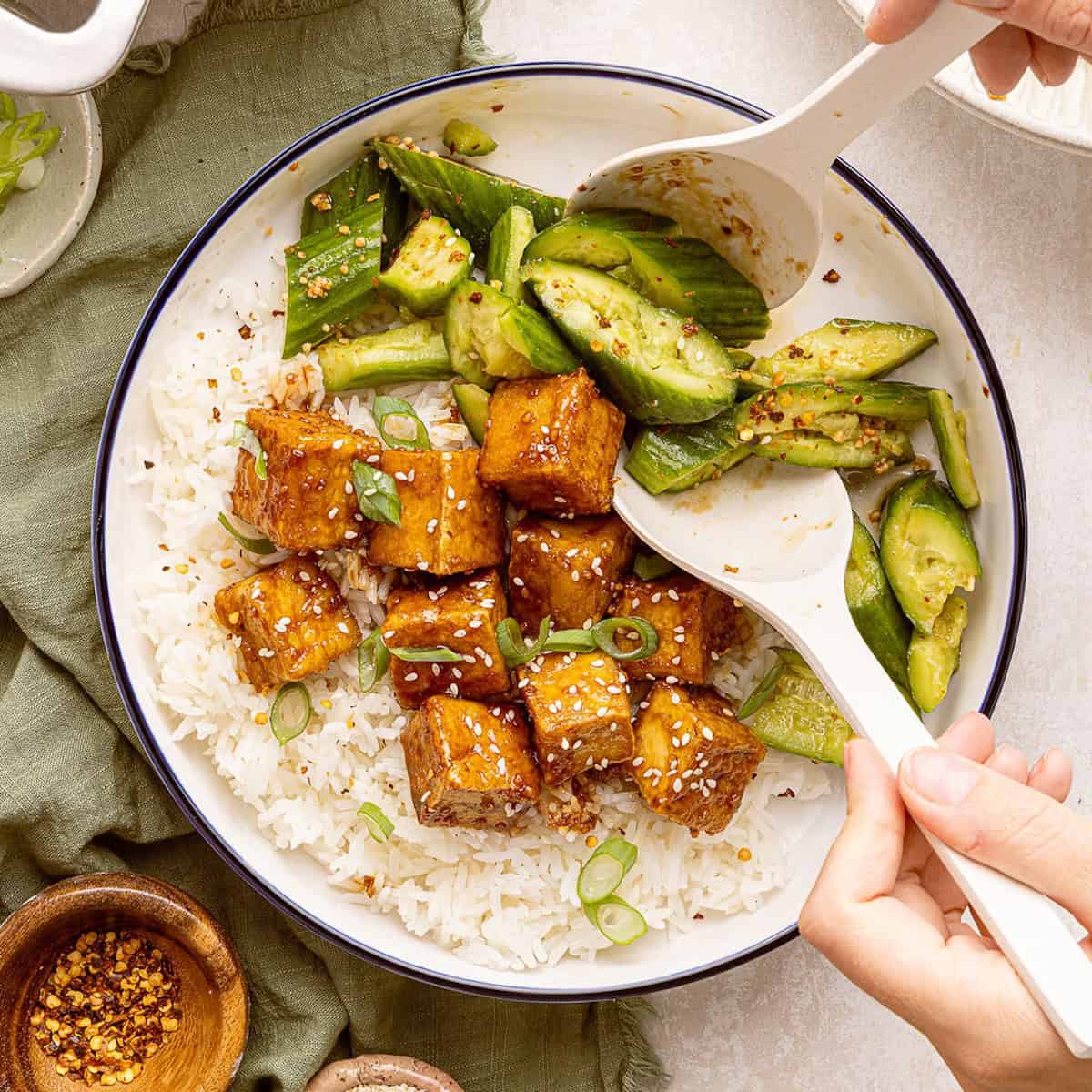 Woman serving smashed cucumber salad next to sticky tofu over rice.