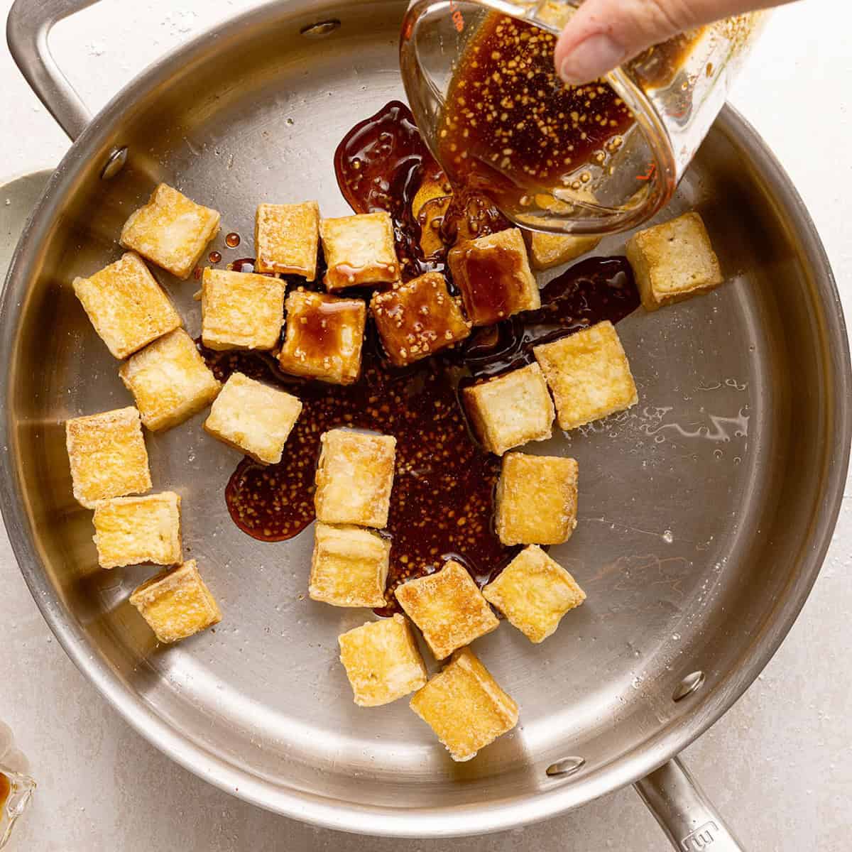 Woman pouring sticky glaze into a skillet with crispy tofu.