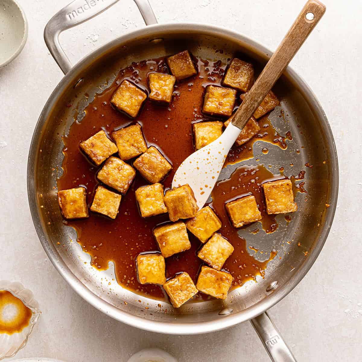 Tofu cooking in a sticky glaze in a skillet until thickened.