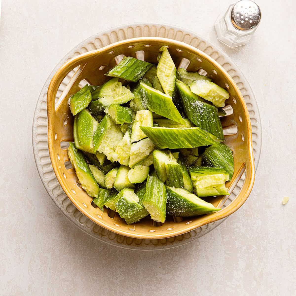 Smashed cucumbers coated in salt and sitting in a colander over a bowl.
