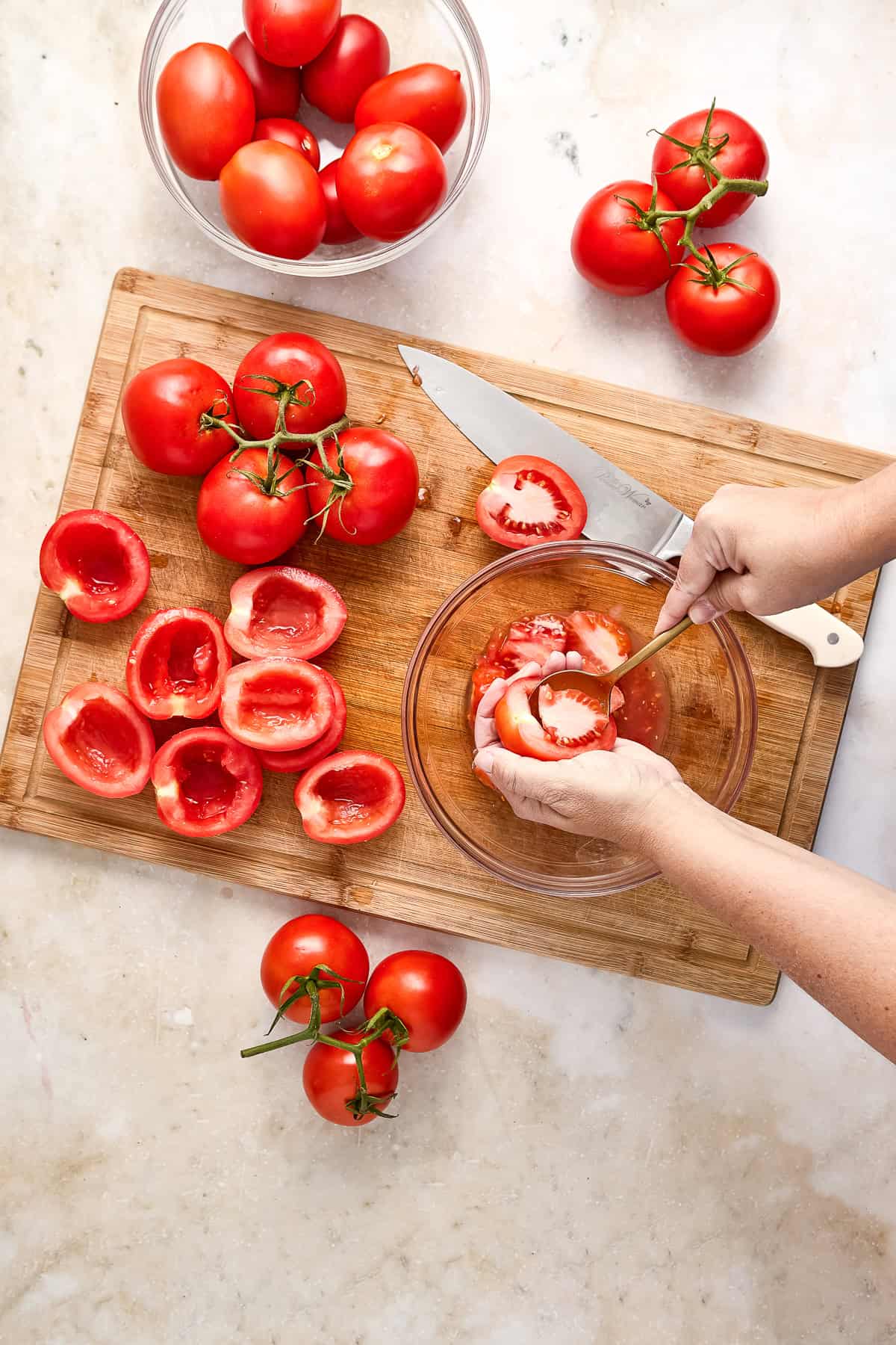Woman scooping seeds and pulp out of tomatoes over a bowl on a cutting board.