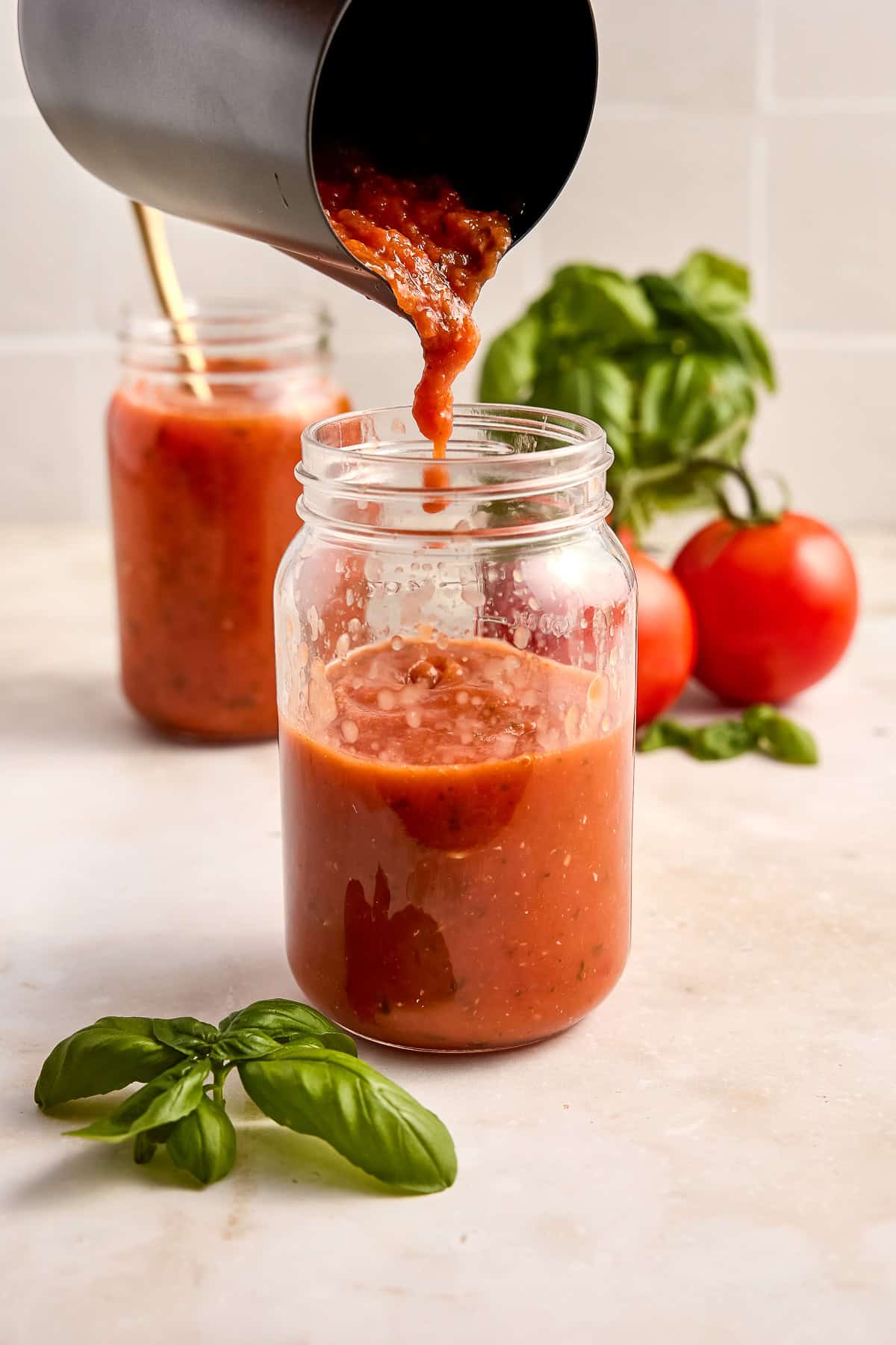 Woman pouring tomato sauce into a jar next to fresh basil and tomatoes.