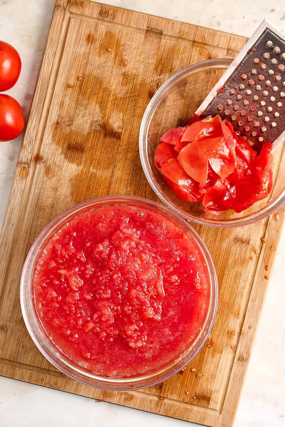 Tomato flesh in a bowl next to another bowl with tomato skins and grater.