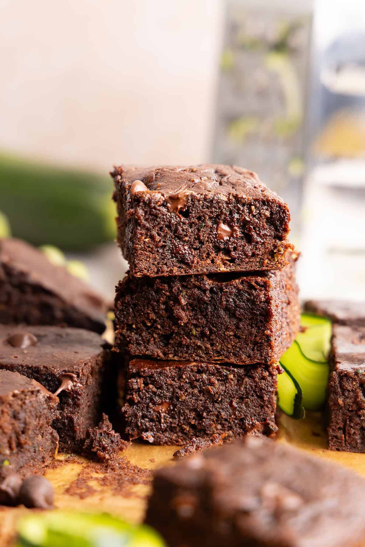 Three zucchini brownies in a stack surrounded by ingredients and more brownies.