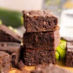 Three zucchini brownies in a stack surrounded by ingredients and more brownies.